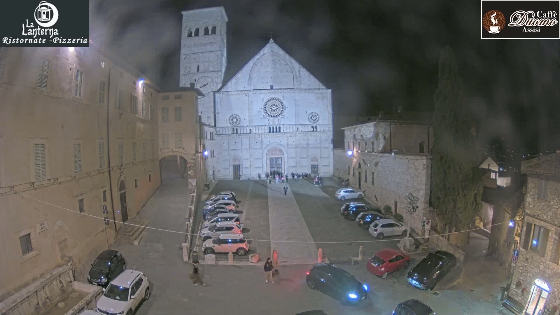 A large, light-colored cathedral stands in a dimly lit town square at night, surrounded by parked cars and buildings, under a hazy sky.