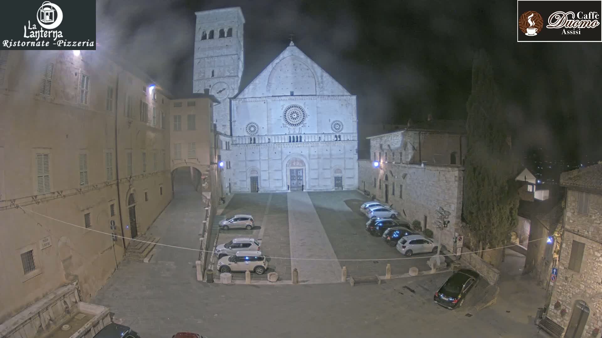 A nighttime view of a town square with parked cars in front of a large cathedral under a hazy sky.