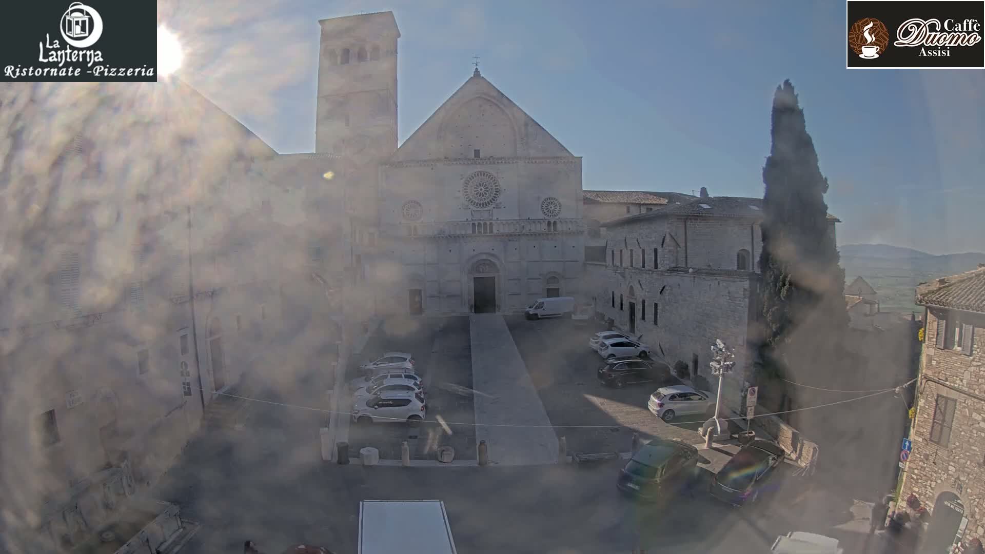 A hazy, sunny day reveals a church courtyard with parked cars, surrounded by stone buildings under a mostly clear sky.