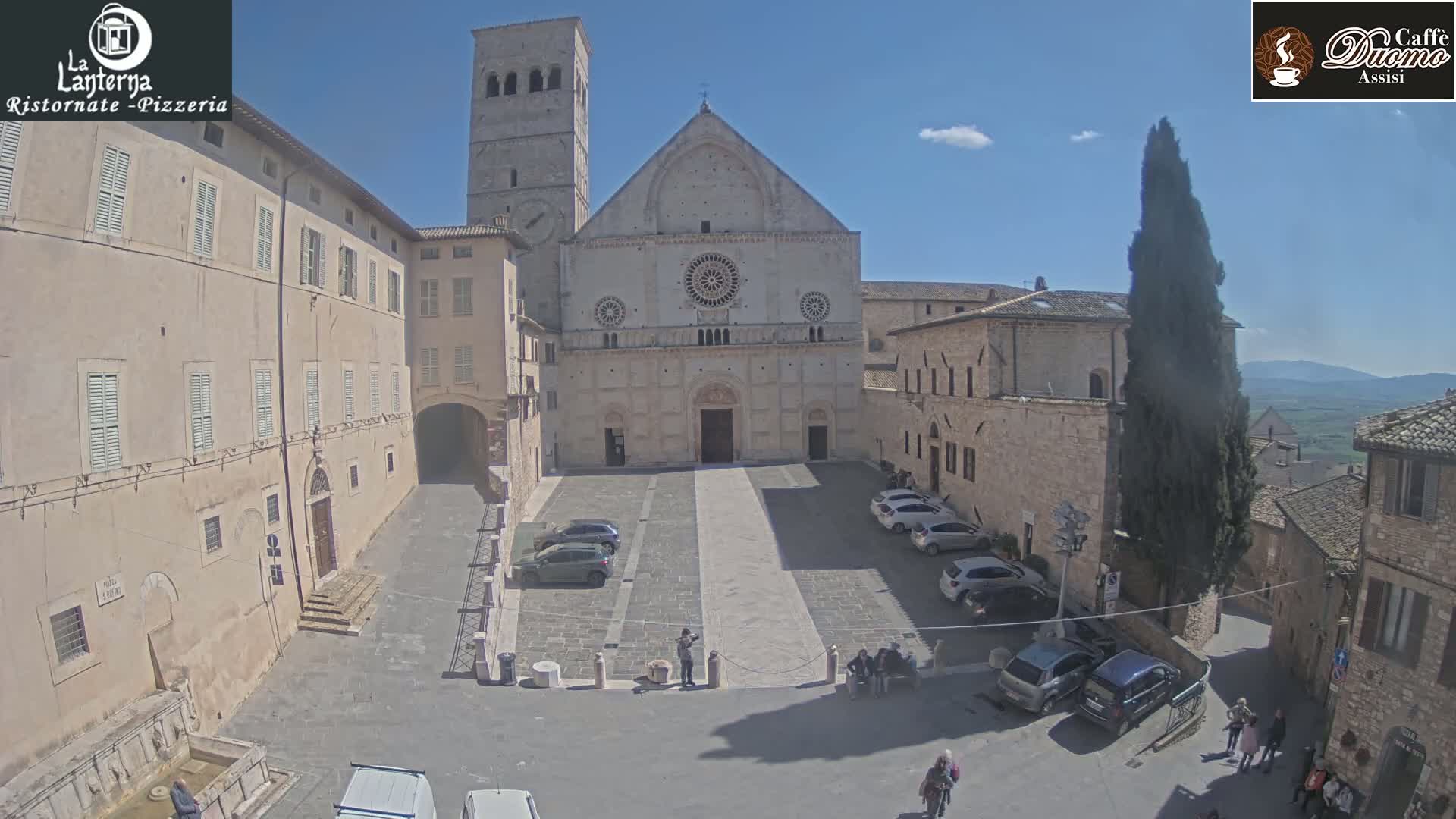 A sunny day reveals a large stone plaza in front of a cathedral, surrounded by buildings, with cars parked and people walking.