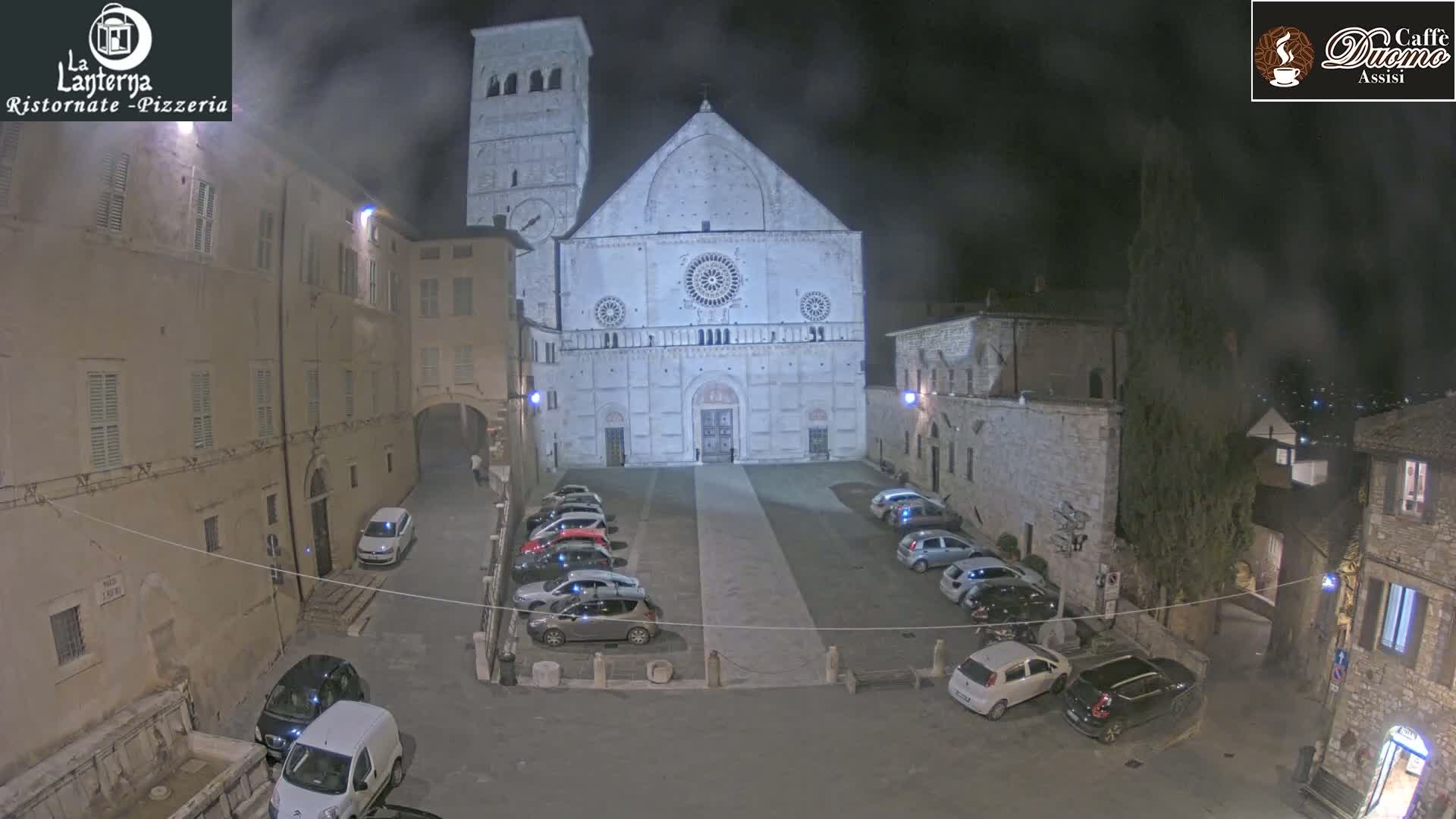 A nighttime view of a town square with a large cathedral at its center, cars parked on either side, and hazy weather conditions.