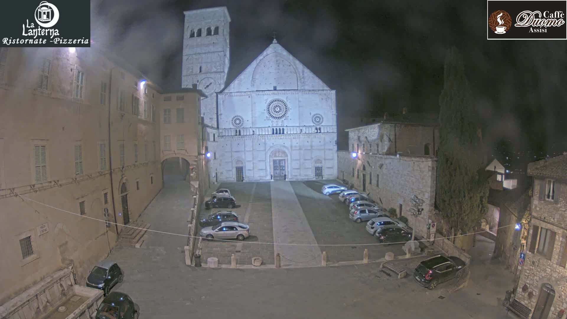 A large cathedral is illuminated at night in a town square with cars parked, under a hazy, dark sky.
