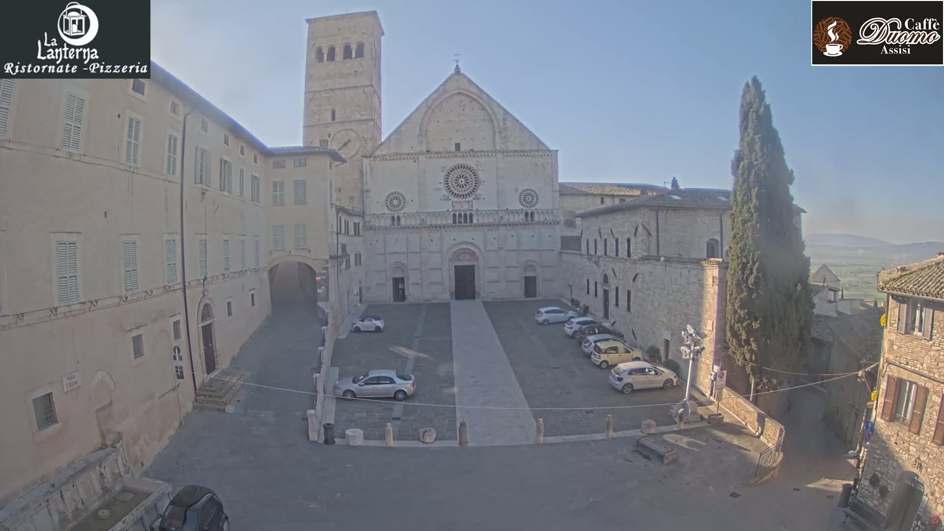 A church with a tall bell tower stands in a town square on a clear, sunny day, with several cars parked in the square.