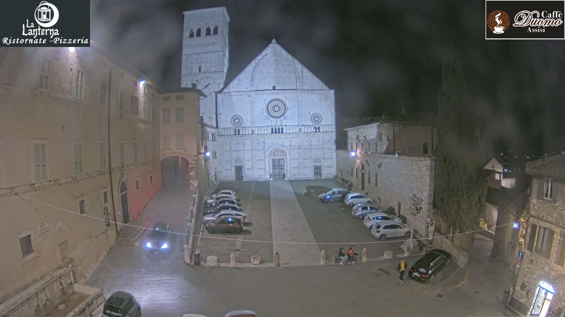 A nighttime view of a town square with a large stone cathedral at its center, several cars parked in the square, and a few people standing near the church.