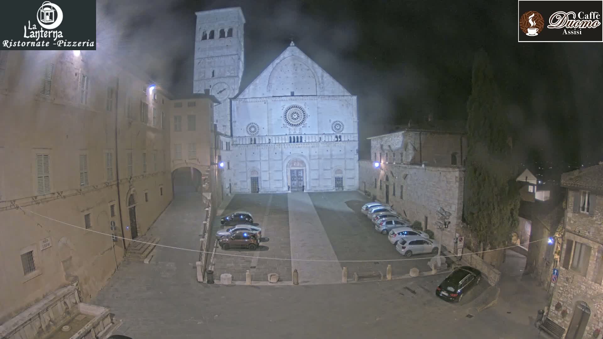 A nighttime view of a town square with a large cathedral at its center, several cars parked in the square, and buildings lining the square under hazy conditions.