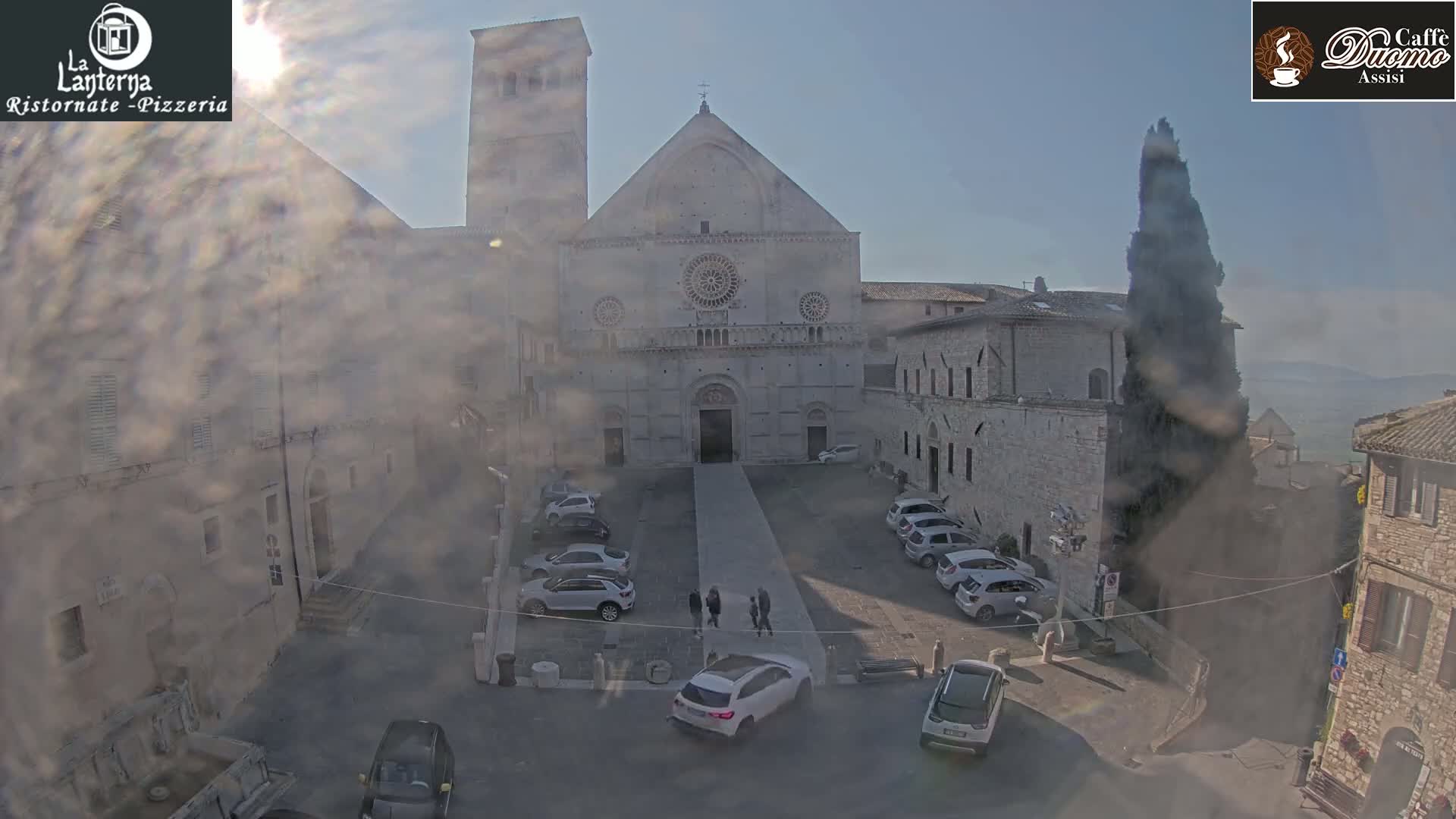 A hazy sun shines on a stone plaza with cars parked around a large cathedral, and several people stand in the plaza.