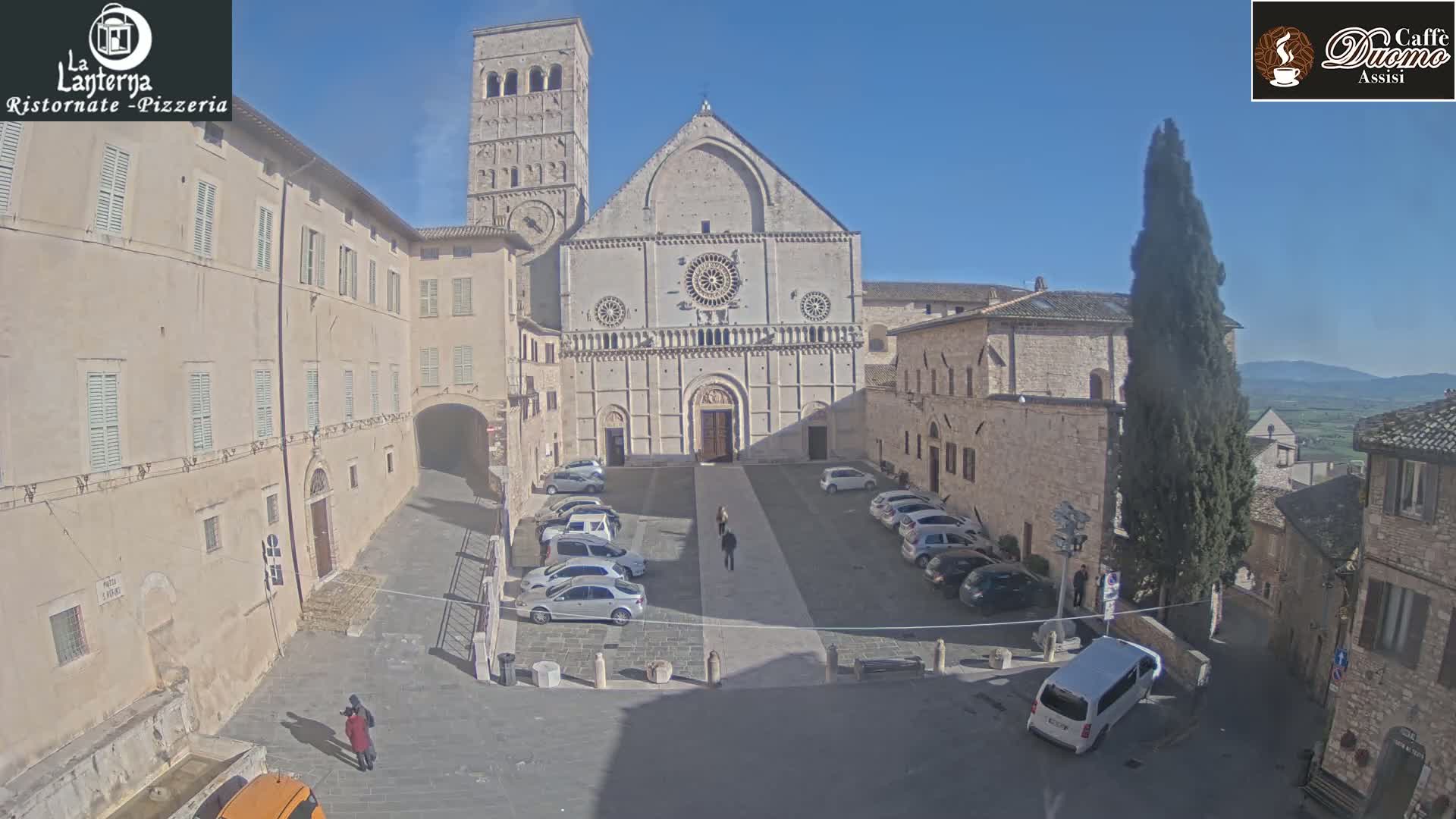 A sunny day reveals a stone plaza with parked cars in front of a large cathedral, surrounded by older stone buildings.