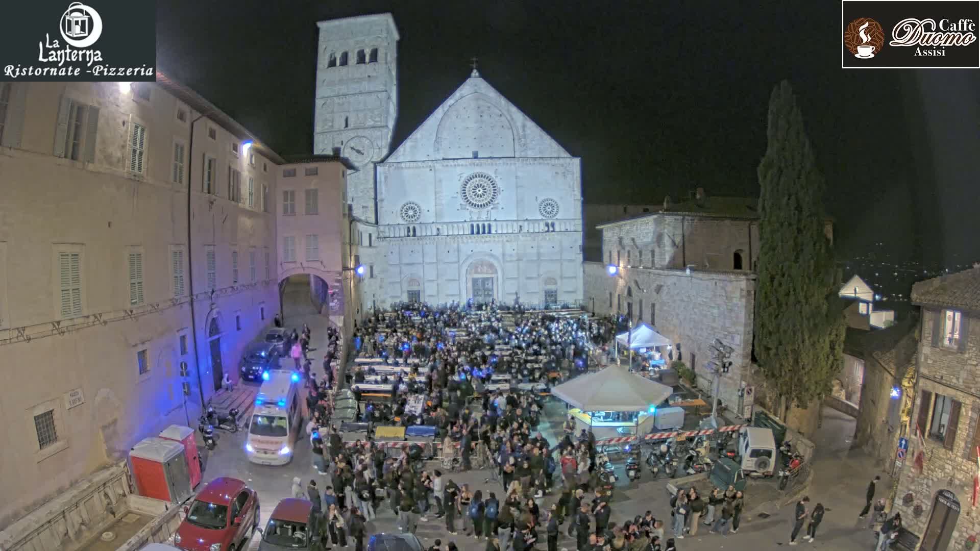 A large crowd gathers in a town square at night in front of a large stone cathedral, under clear skies.