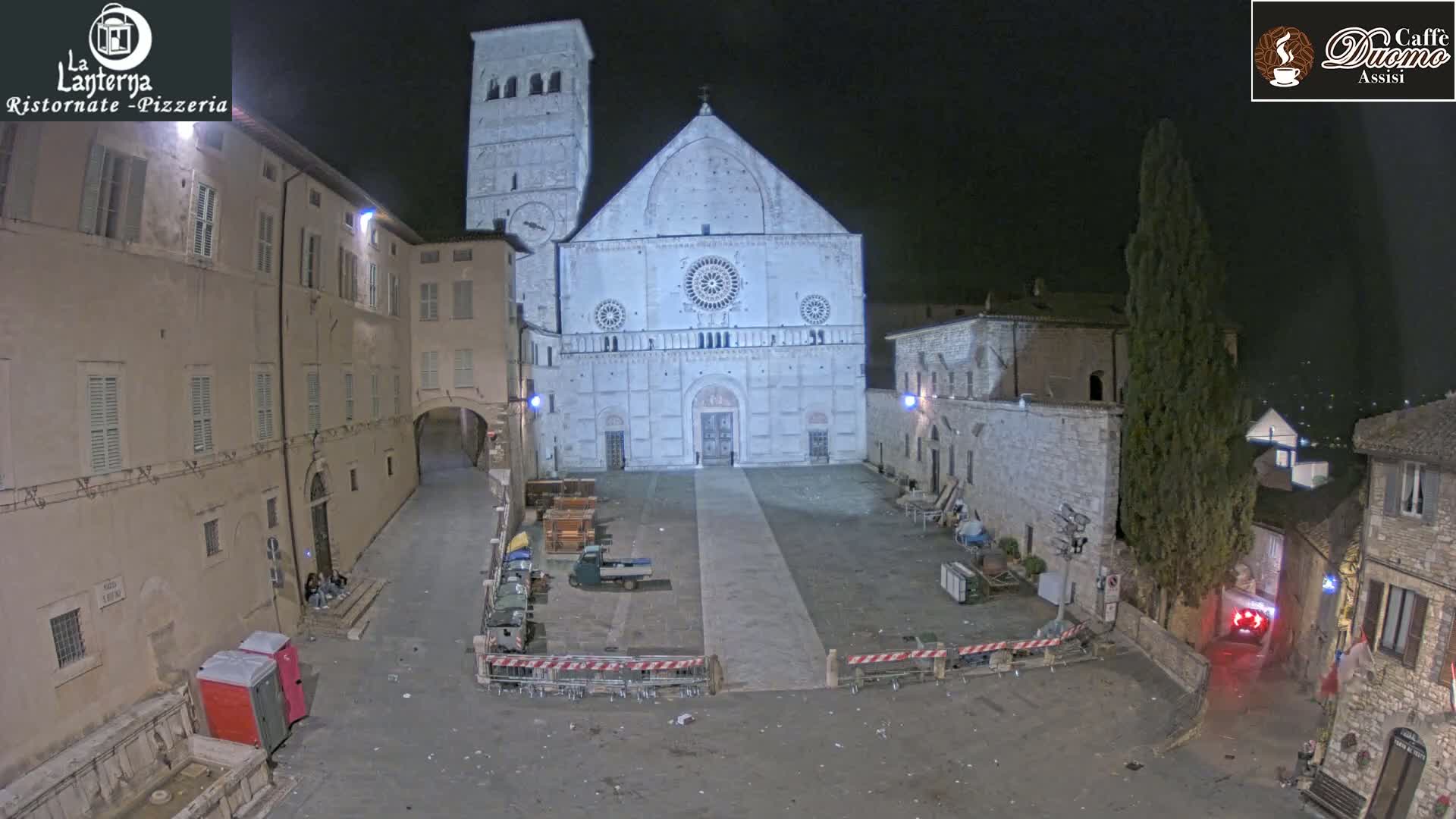 At night, a large, light-colored cathedral stands in a town square surrounded by stone buildings.
