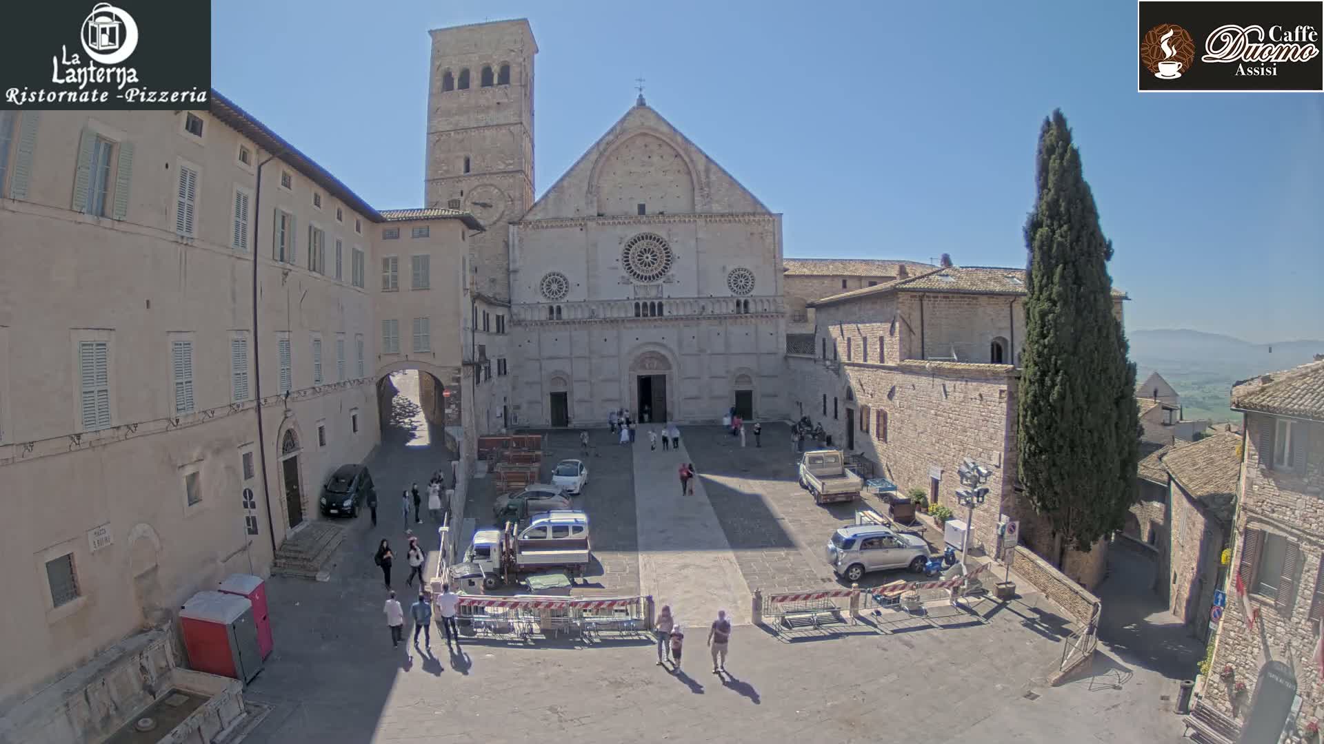 A large stone cathedral stands in a sunny plaza, surrounded by low buildings, parked vehicles, and a few pedestrians.