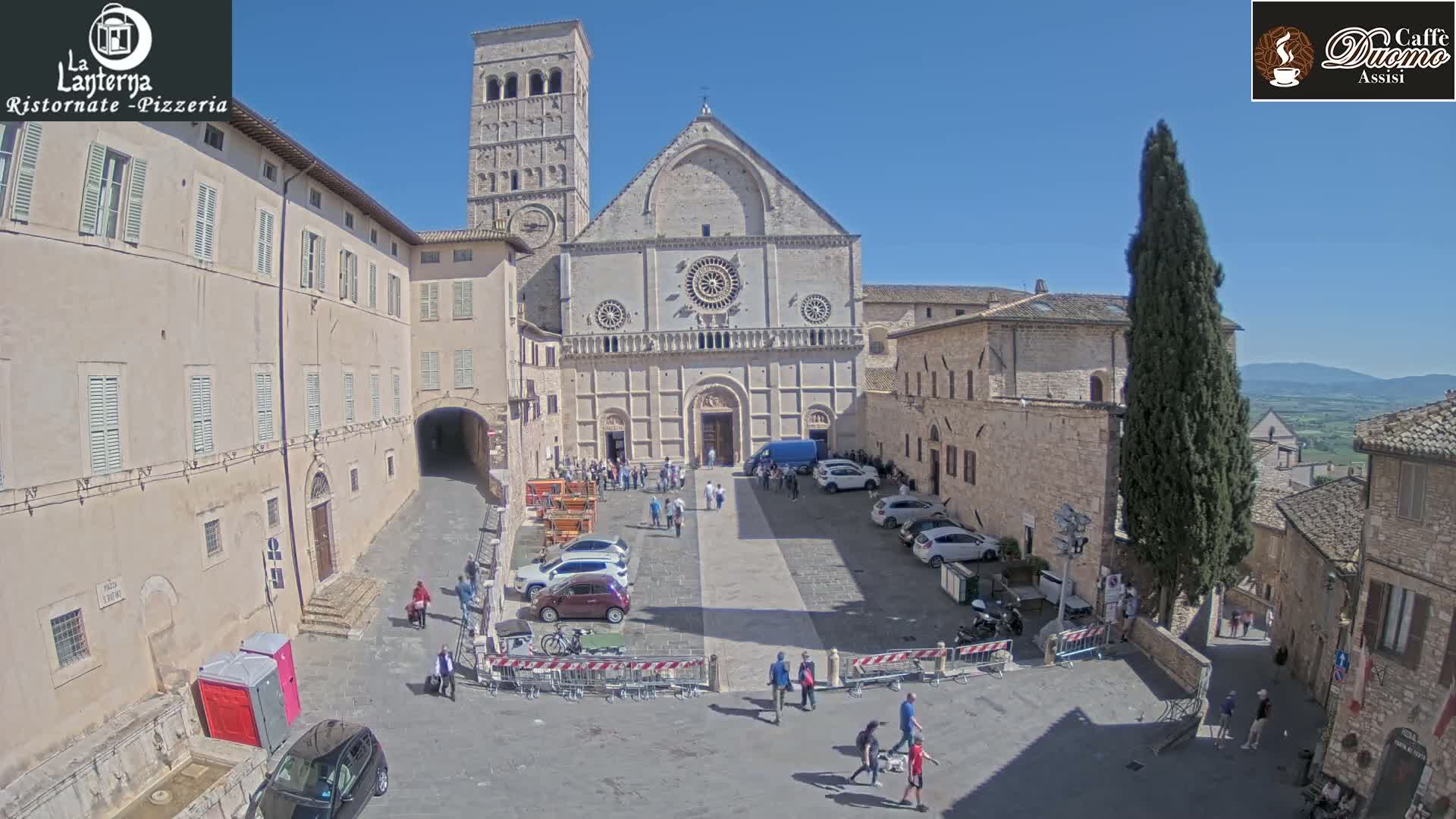A large stone church and surrounding buildings are visible in a sunny, outdoor square with several parked cars and pedestrians.