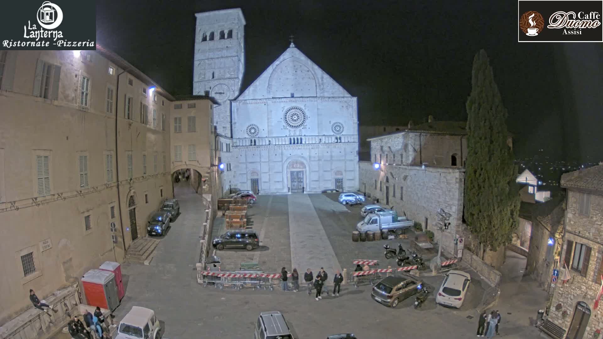 A nighttime aerial view of a town square shows a large cathedral, several parked cars, and a few pedestrians, under a clear sky.