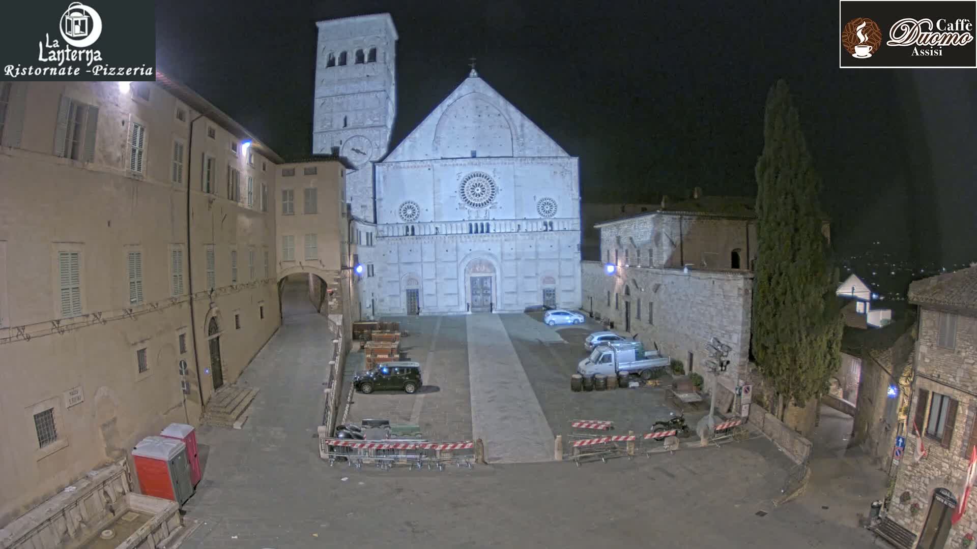 A large stone cathedral is visible at night in a town square, with several vehicles parked nearby and some barriers in place.