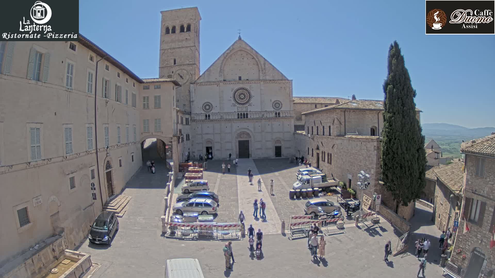 A sunny outdoor scene shows a large stone church surrounded by buildings and parked cars in a town square with several people walking around.