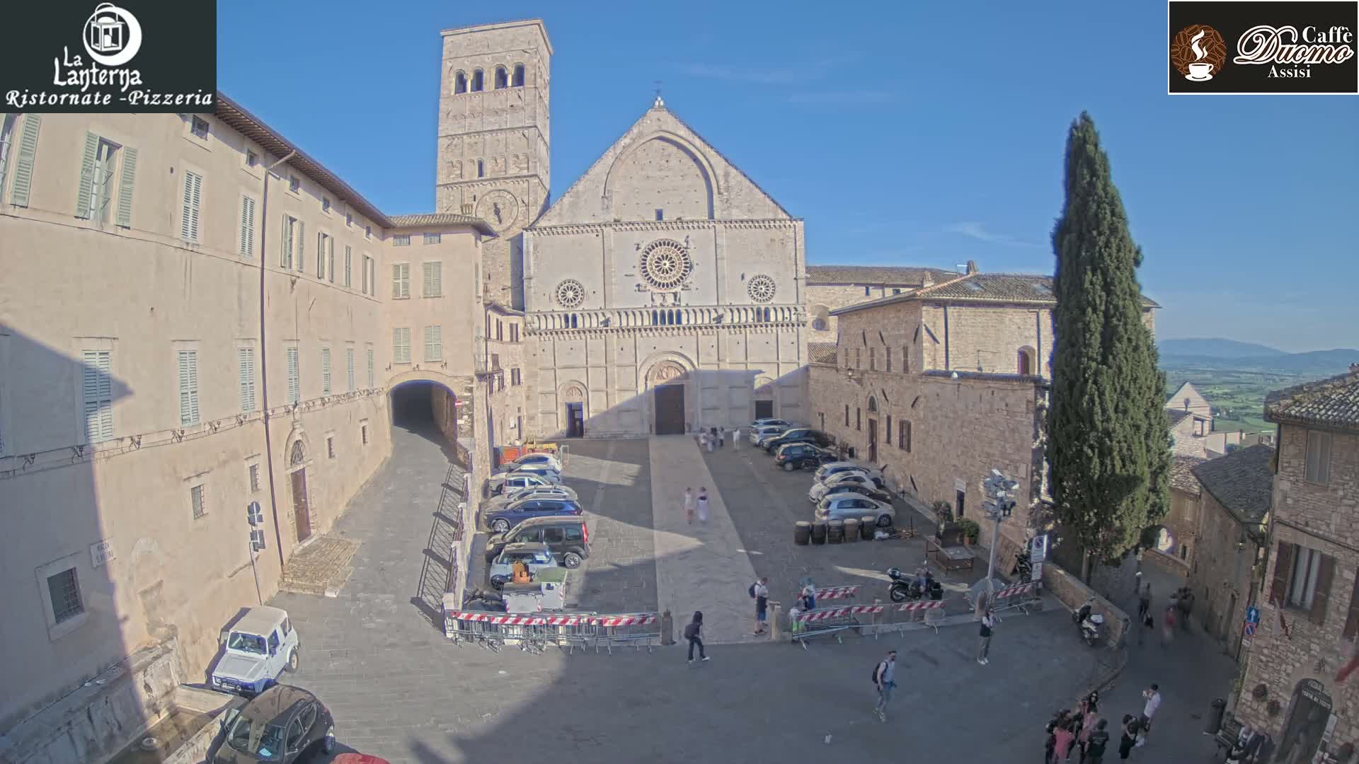 A large stone cathedral sits in a sunny town square surrounded by buildings and parked cars.