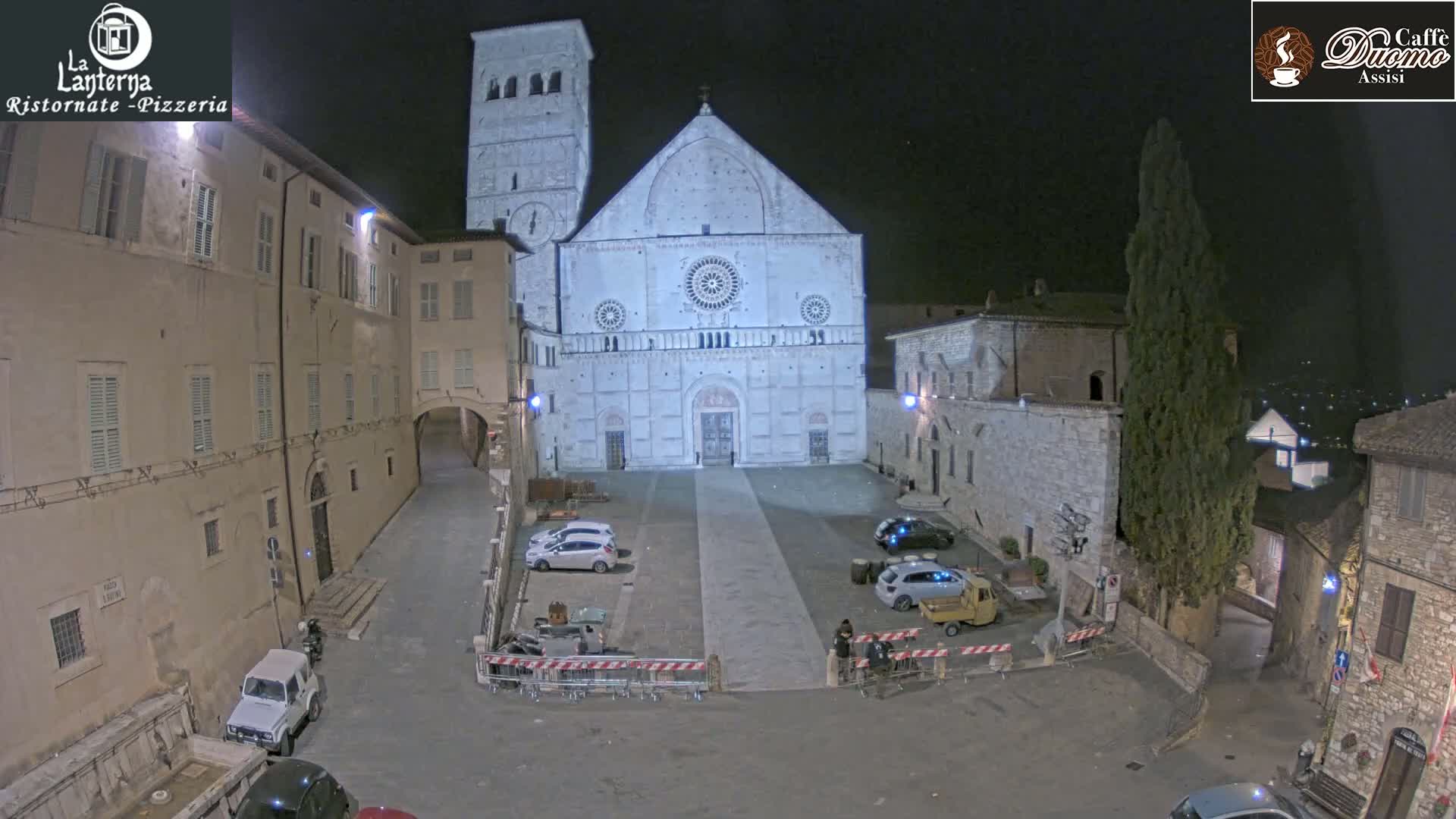 A large stone cathedral stands in a town square at night, with several cars parked and a few people visible under artificial light.