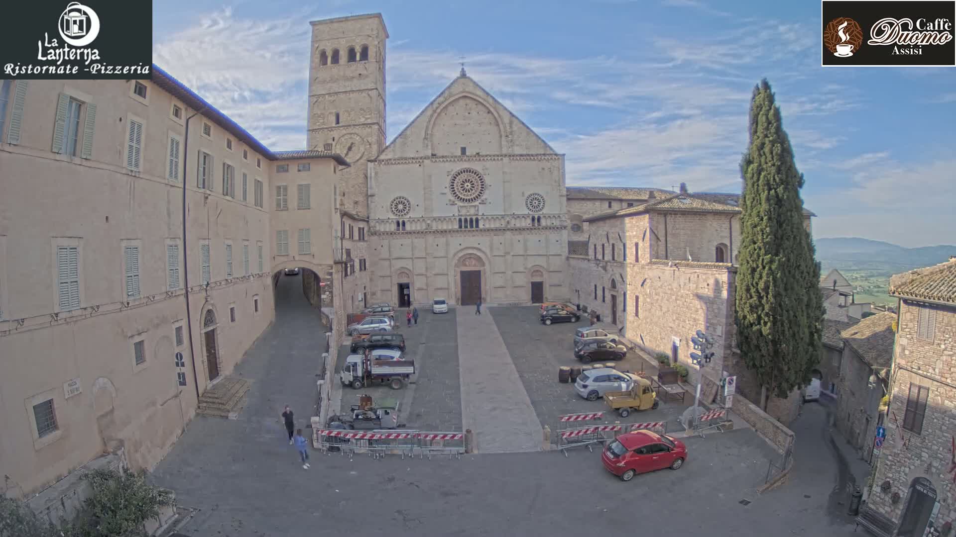 A stone cathedral and surrounding buildings are visible in a town square under a mostly clear sky, with a few cars parked and some people walking nearby.