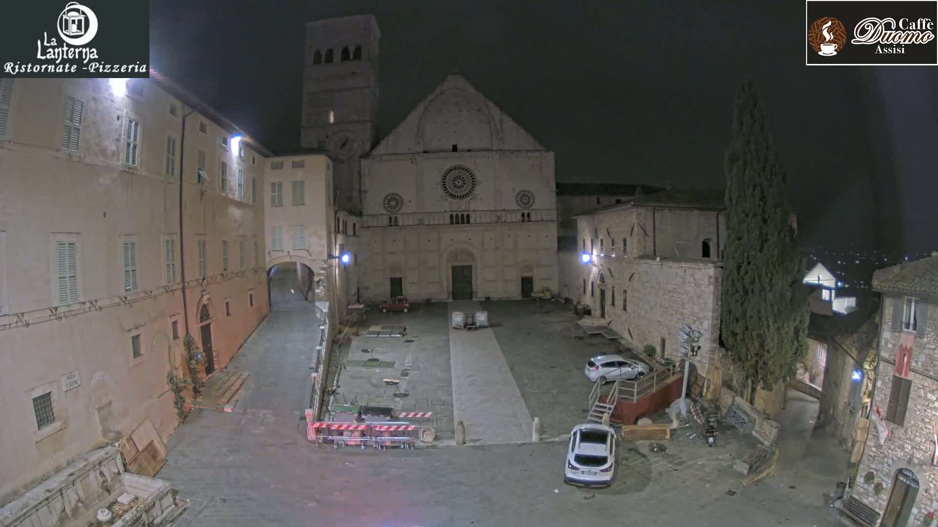 A nighttime view of a large, stone plaza in front of a cathedral, with several cars parked and some construction materials visible.