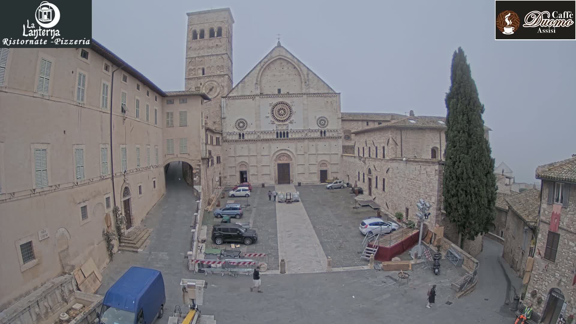A stone cathedral and surrounding buildings are visible in a town square on an overcast day, with several cars parked and some construction equipment present.