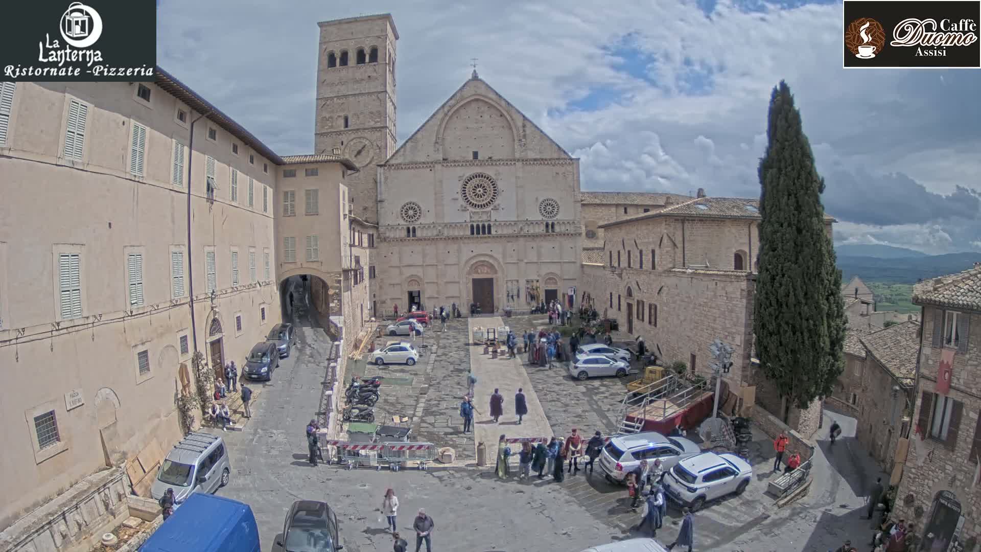 A large stone cathedral and surrounding buildings, with people gathered in the square, under partly cloudy skies.