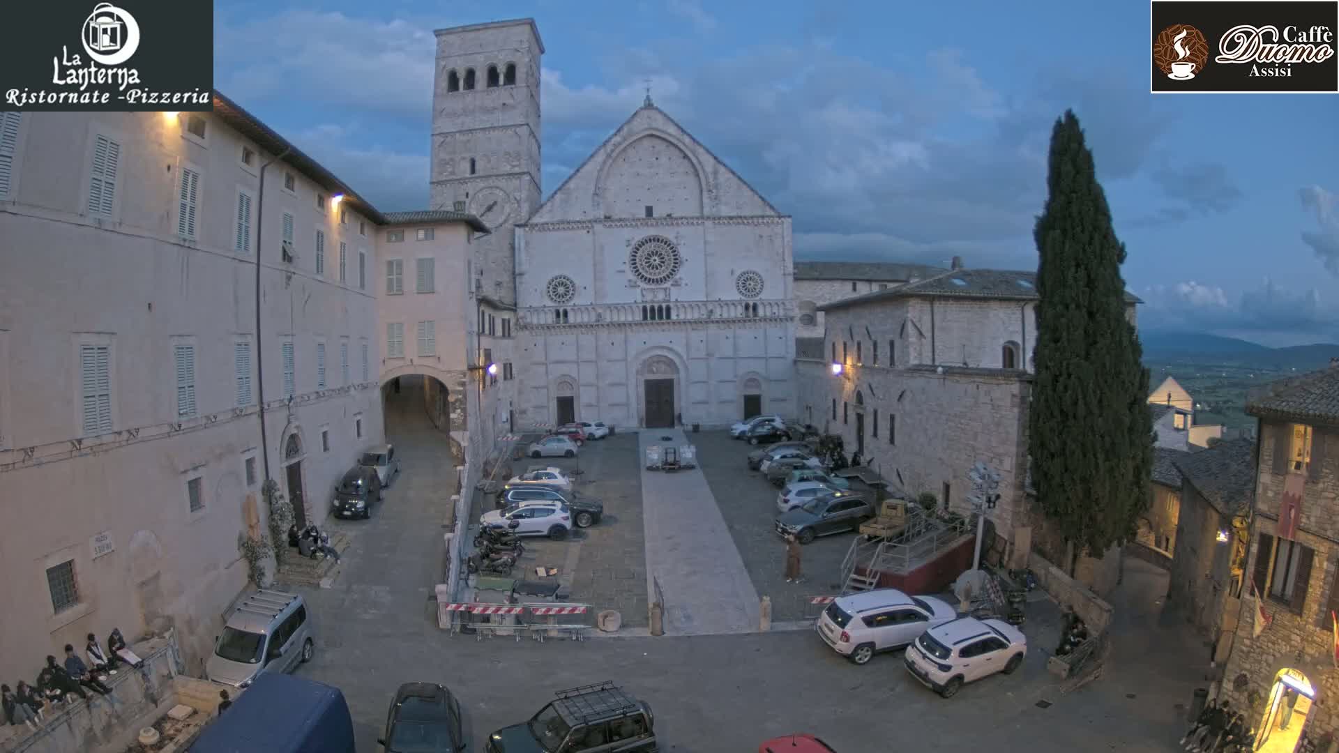 A large stone cathedral stands in a town square at dusk, surrounded by buildings and parked cars under a cloudy sky.