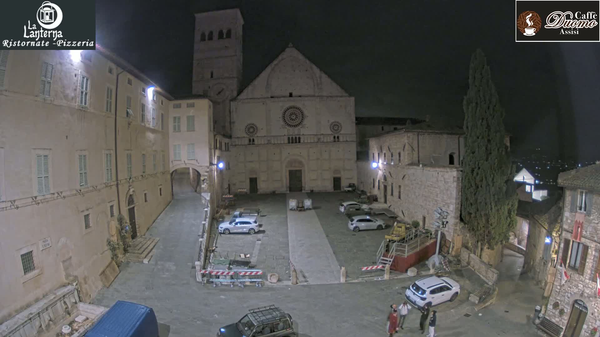 A nighttime, aerial view of a town square in front of a large stone church, with several cars parked and a few people standing near some construction barriers.