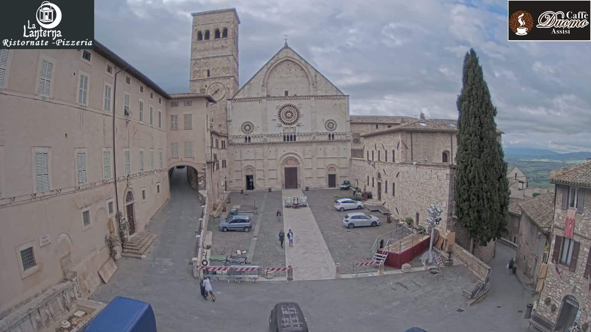 A large stone cathedral stands in a town square under an overcast sky, surrounded by older stone buildings, cars, and a few pedestrians.