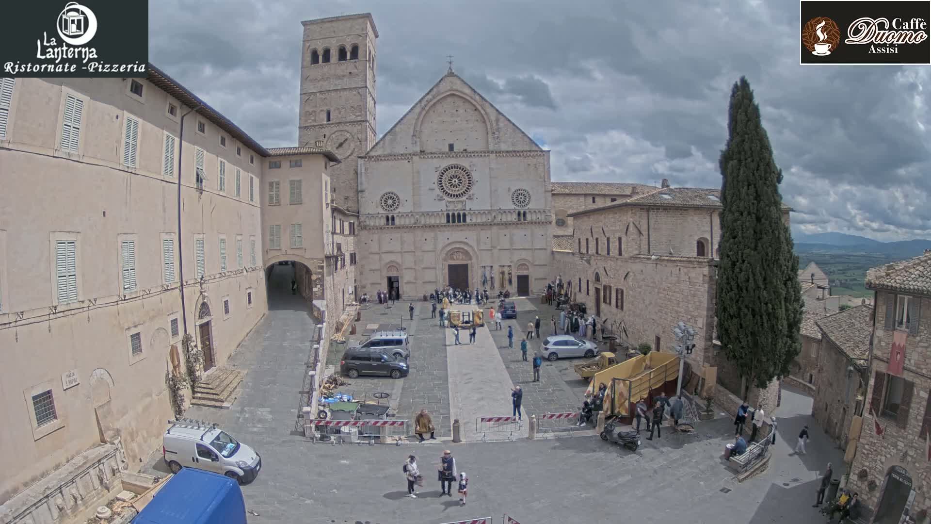 A large stone cathedral stands in a town square under a cloudy sky, surrounded by other stone buildings and several people.
