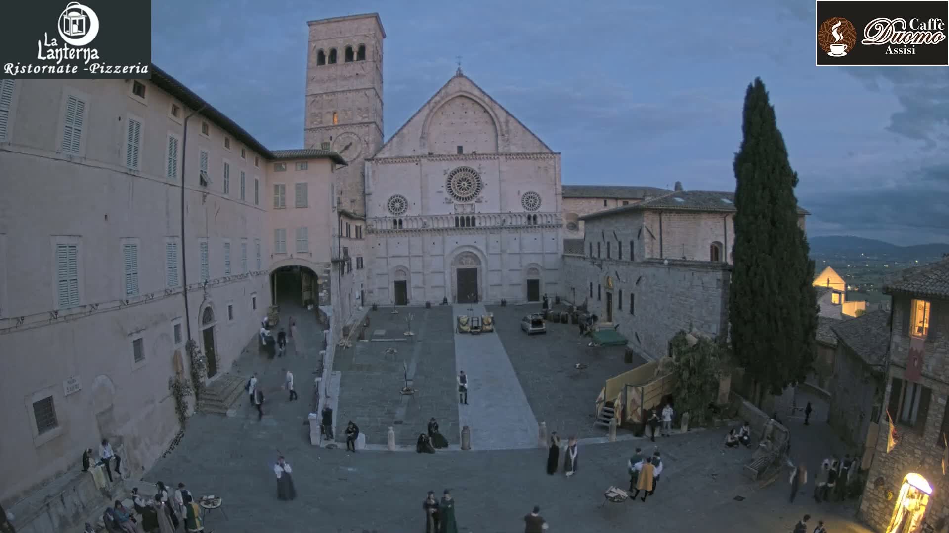 A large stone cathedral and surrounding buildings are seen in a town square under a twilight sky, with many people in medieval-style clothing gathered in the plaza.