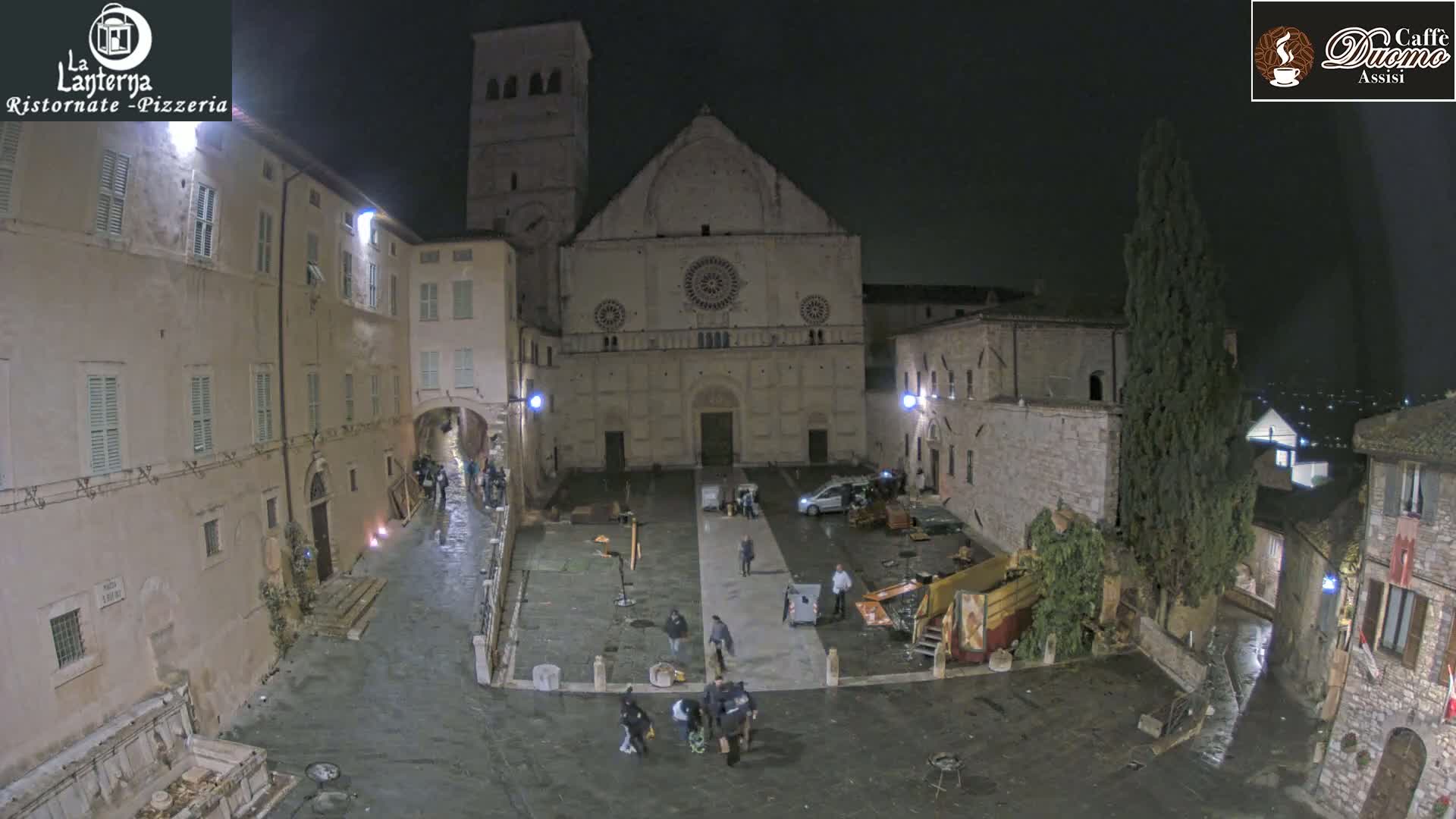 A nighttime, wet town square is shown, centered on a large, light-colored stone building with several people and vehicles present.