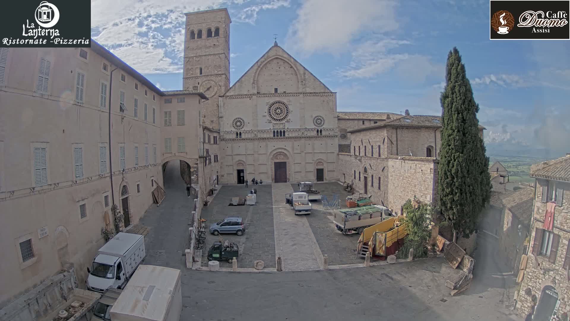 A large stone cathedral stands in a town square on a partly cloudy day, surrounded by old buildings and several vehicles.
