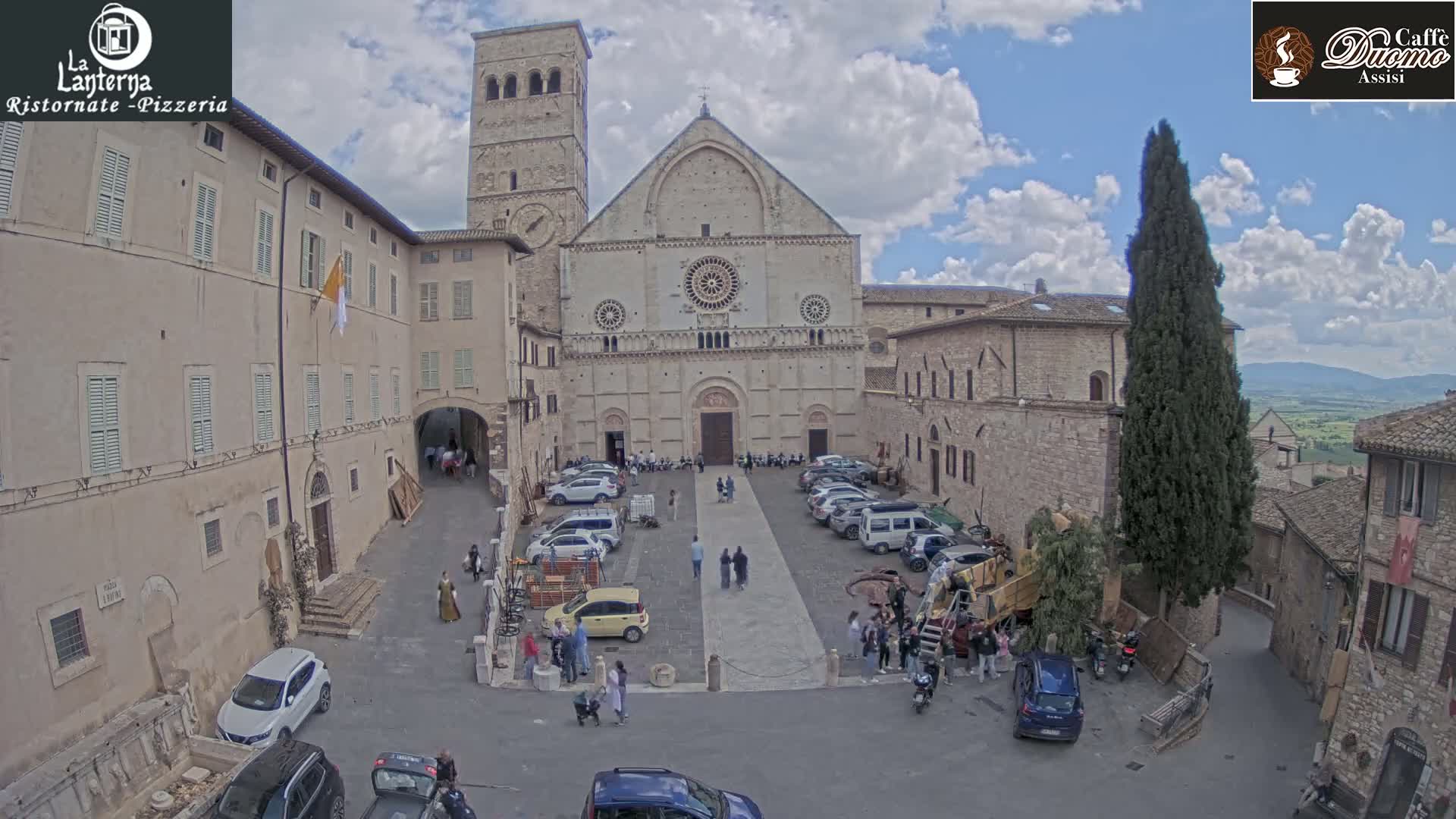 A large stone cathedral stands in a town square on a partly cloudy day, surrounded by other stone buildings and parked cars, with several people walking around.
