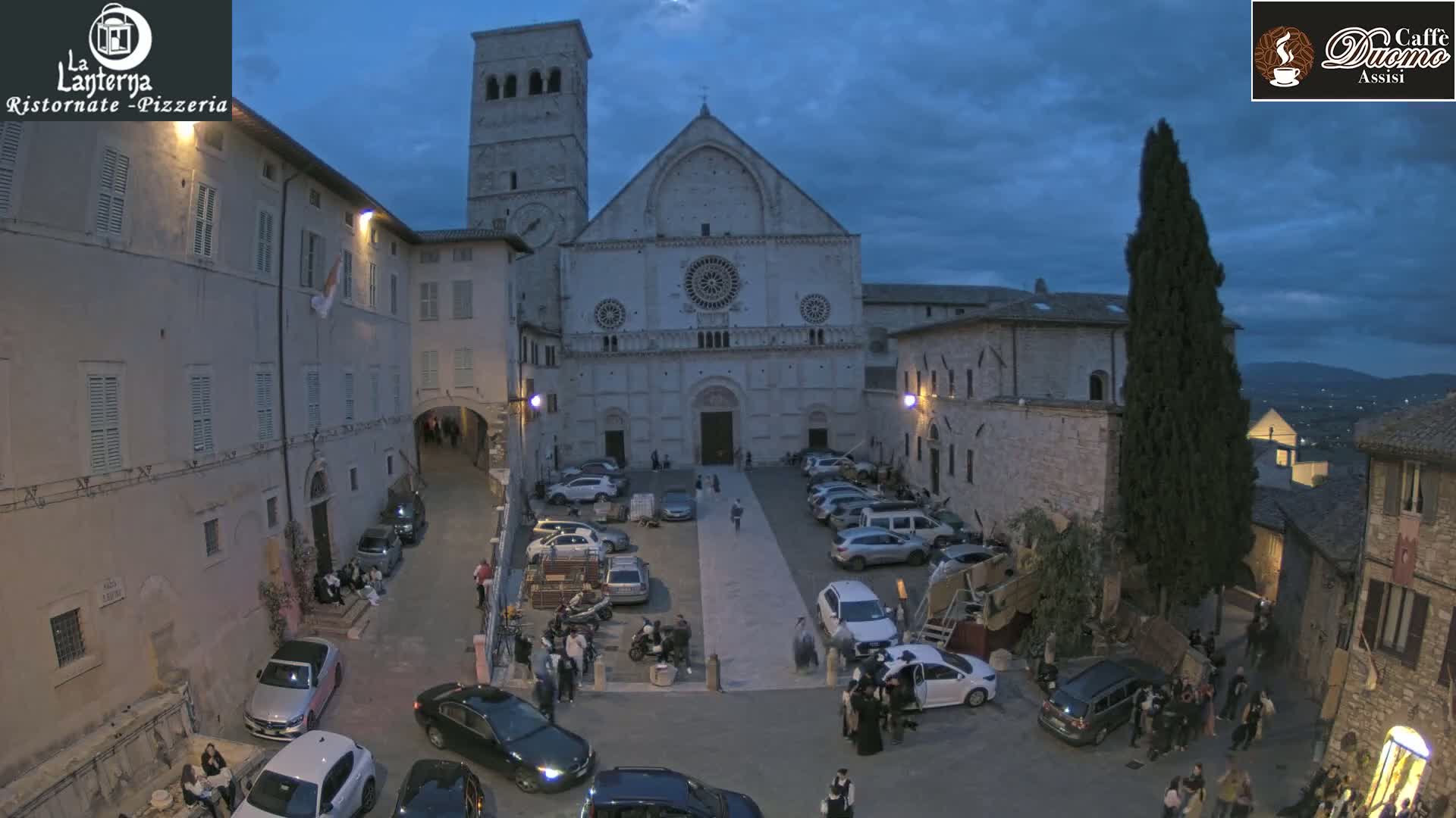 A large stone cathedral is situated in a town square at dusk, surrounded by buildings and parked cars under a cloudy sky.