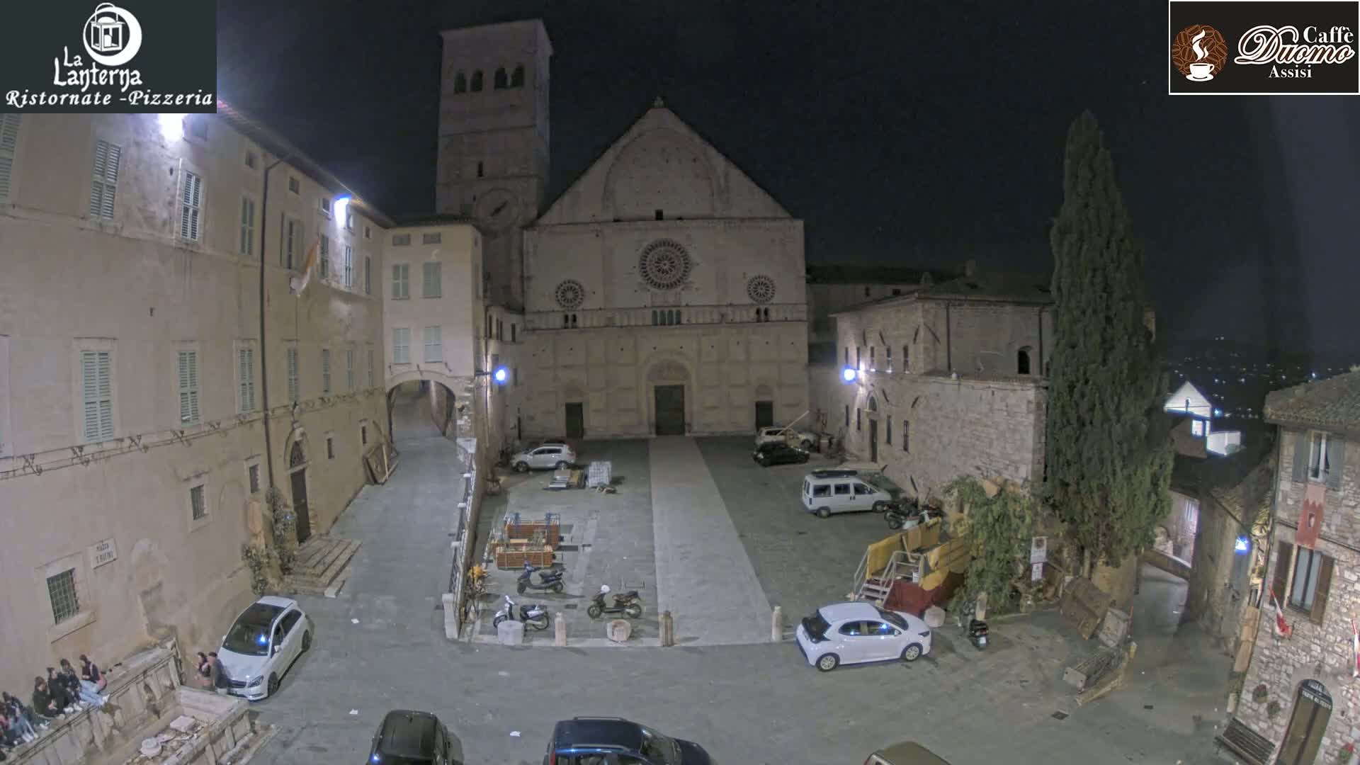 A nighttime view of a town square dominated by a large stone church, with several parked cars and a few pedestrians visible under a clear, dark sky.