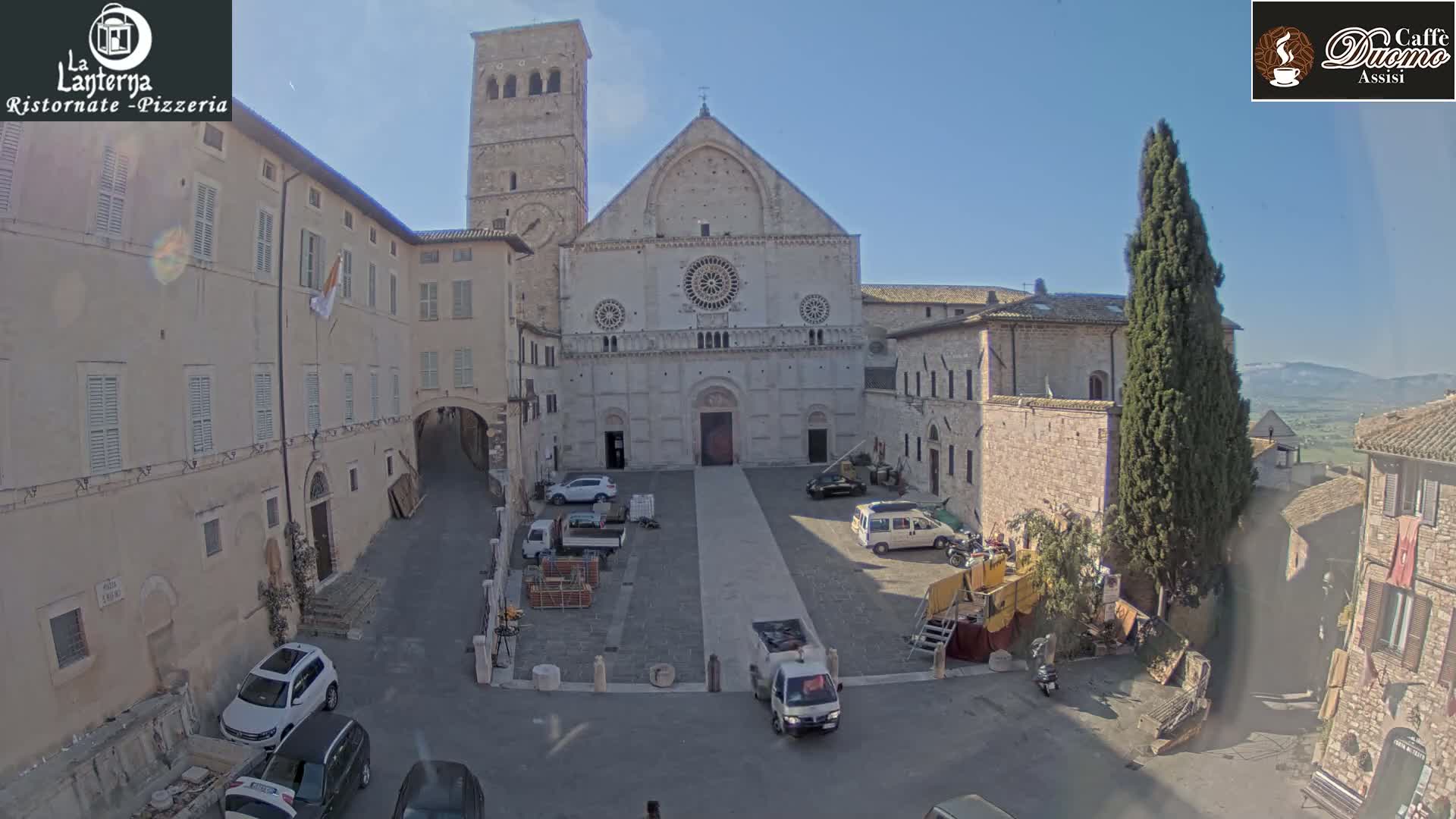 A large stone cathedral stands in a plaza on a sunny day, surrounded by older stone buildings, several vehicles, and construction materials.