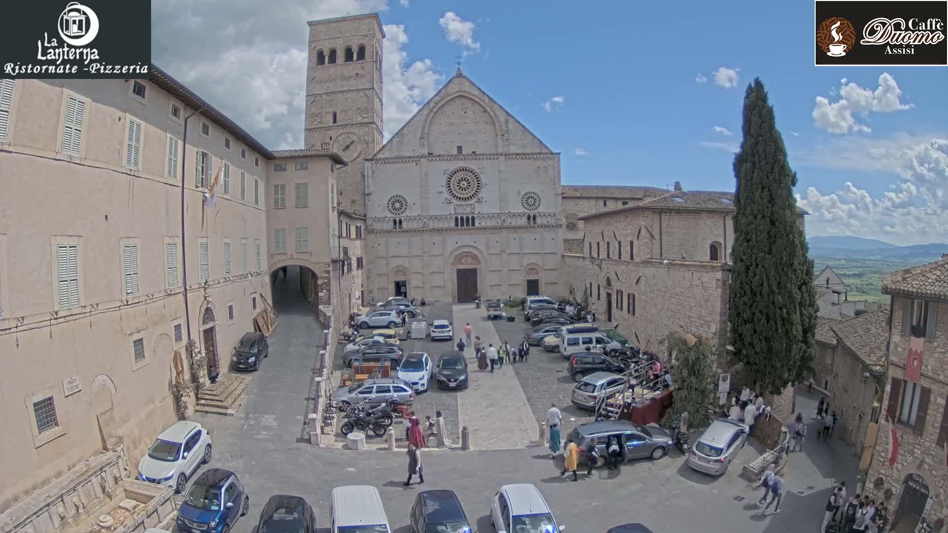 A town square with a large stone church in the background, surrounded by buildings and parked cars under a partly sunny sky.