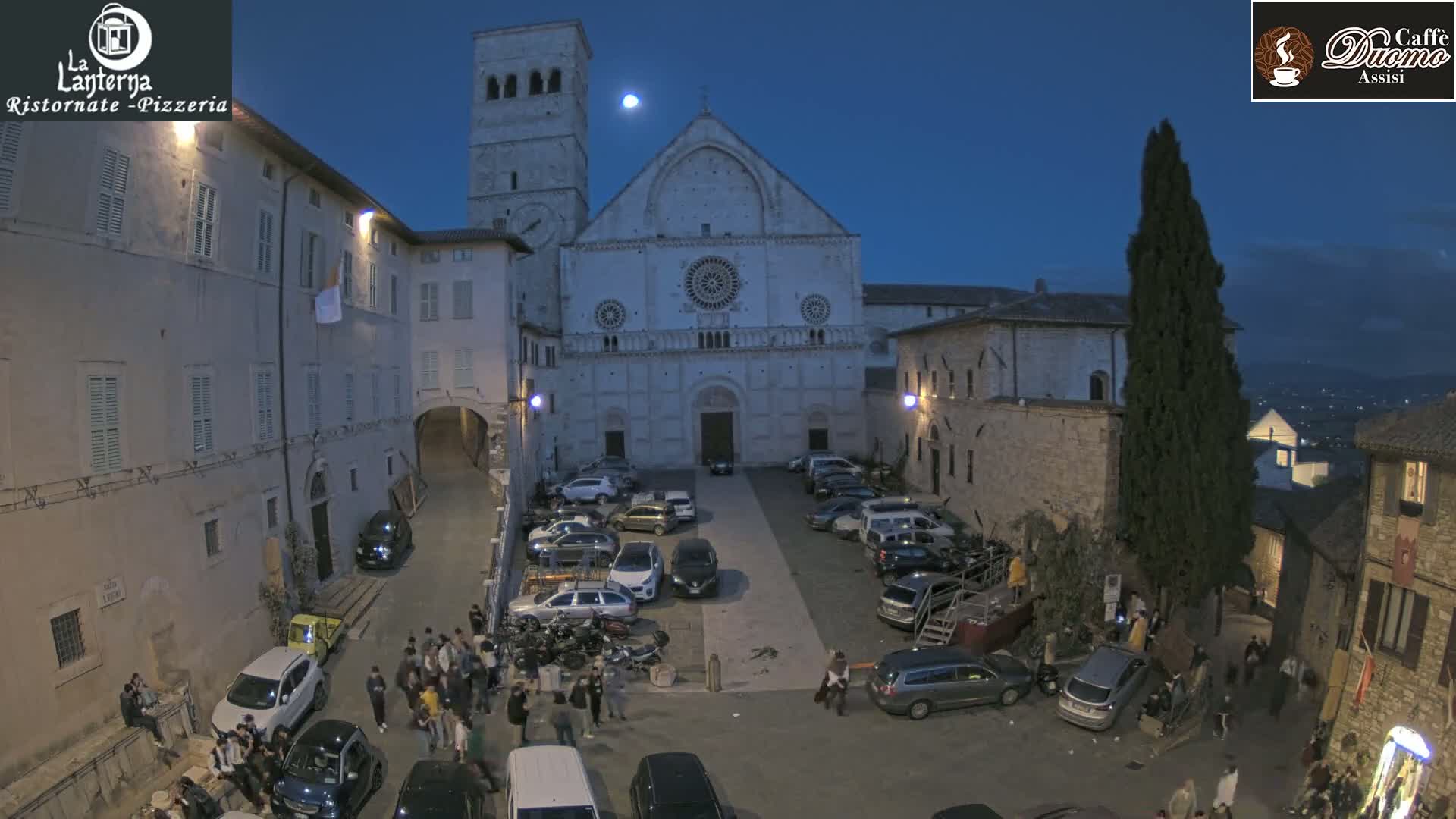 A town square at twilight, under a partly cloudy night sky with a visible moon, is filled with parked cars and people gathered around the base of a large stone cathedral.