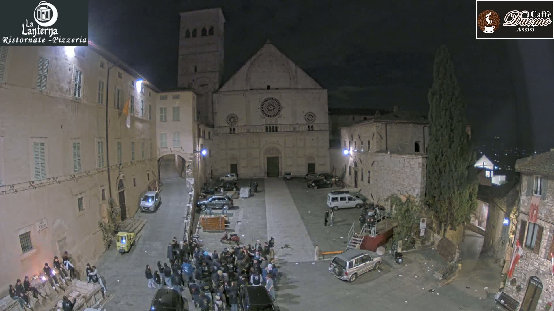 A nighttime view of a town square with a large stone church at its center, surrounded by buildings and a crowd of people gathered in the square.