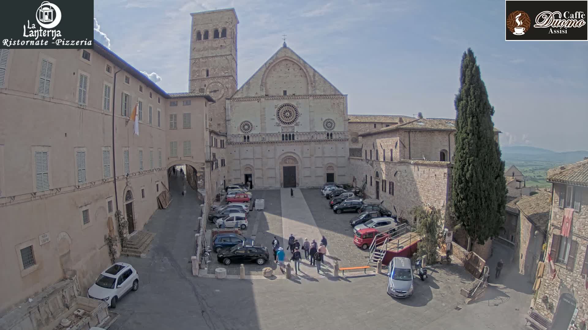 A large stone church stands in a sunny town square, surrounded by buildings and parked cars, with a few people walking in the plaza.