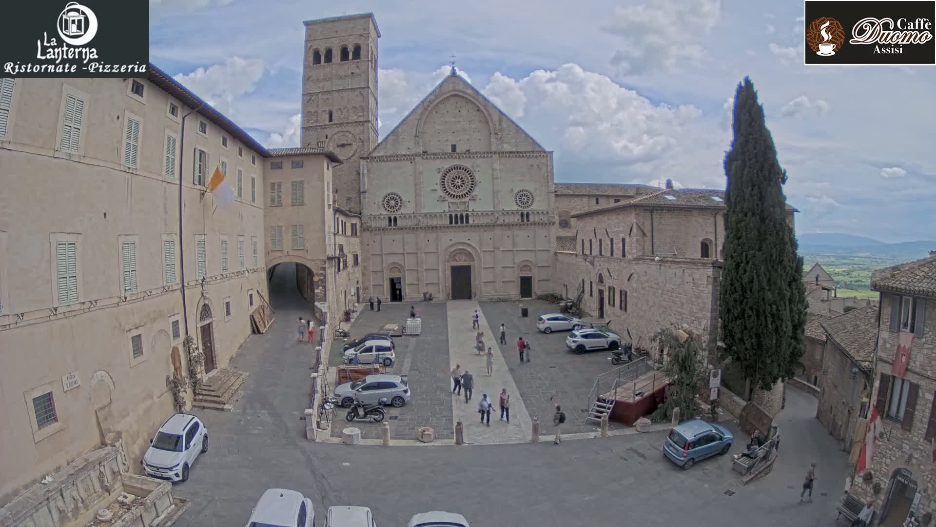 A large stone church faces a town square under a partly cloudy sky, with several cars and people in the square and surrounding buildings.
