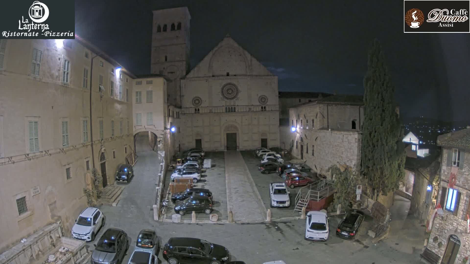 A nighttime, high-angle view of a town square with parked cars in front of a large stone church and surrounding buildings.