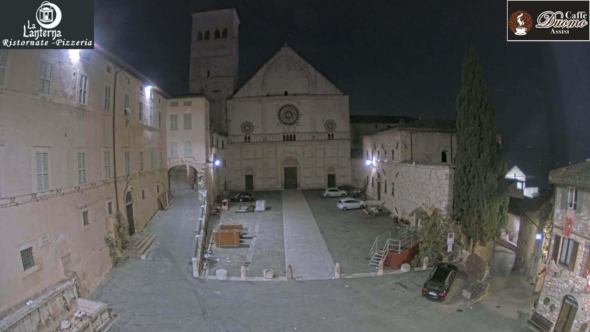 At night, a mostly empty town square is seen in front of a large stone church, with a few cars parked and some buildings surrounding the square.