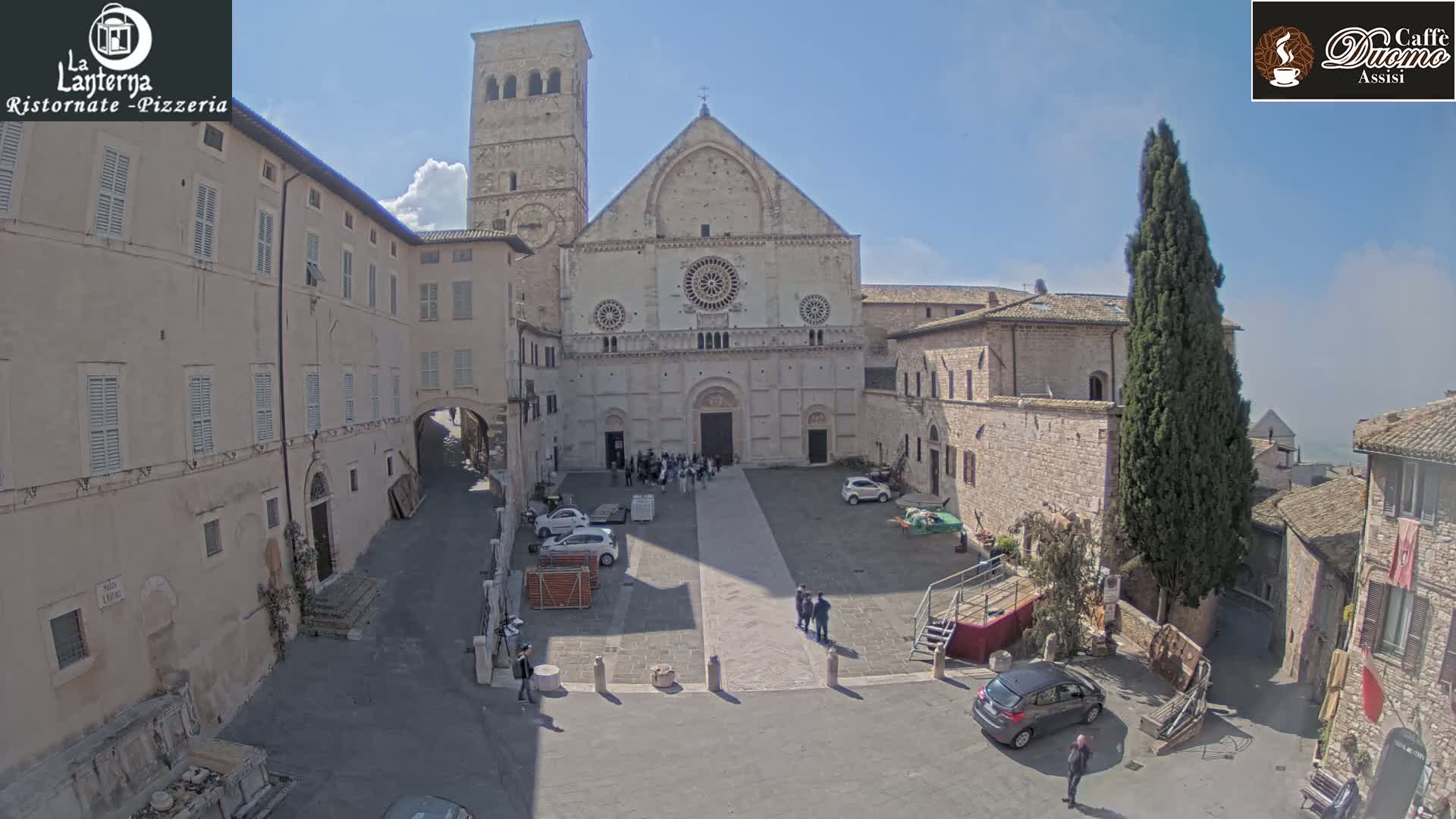 A large stone cathedral stands in a town square on a partly sunny day, surrounded by other stone buildings and a few parked cars.