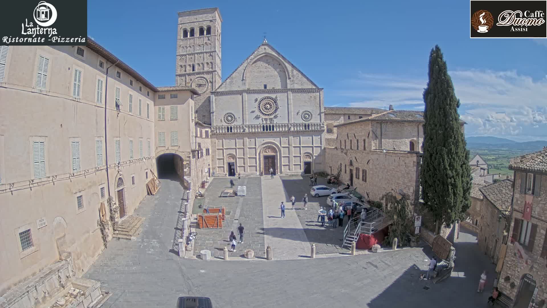 A sunny day reveals a town square with a large stone cathedral as its centerpiece, flanked by aged buildings, and a few people scattered throughout.