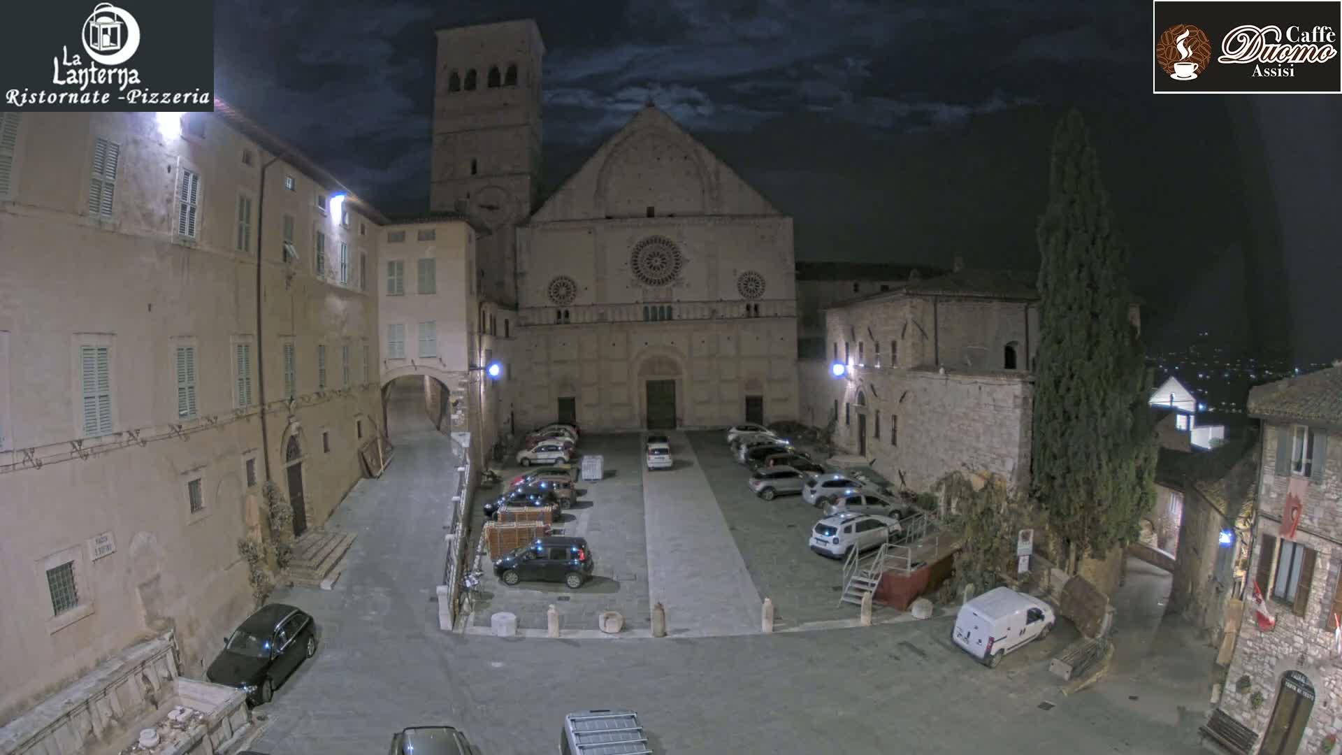 A nighttime view of a stone plaza with parked cars in front of a large stone church under a partly cloudy sky.