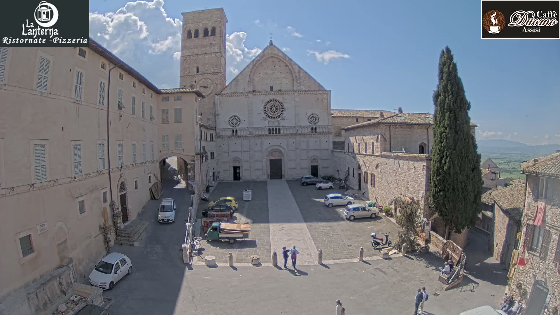 A large stone cathedral sits in a town square under a partly cloudy sky, surrounded by buildings and a few cars and people.