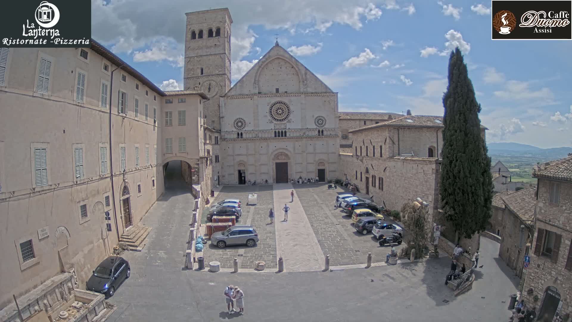 A large, light-colored stone church with a rose window and bell tower is situated in a town square under a partly sunny sky; several cars are parked in the square, and a few people are walking around.