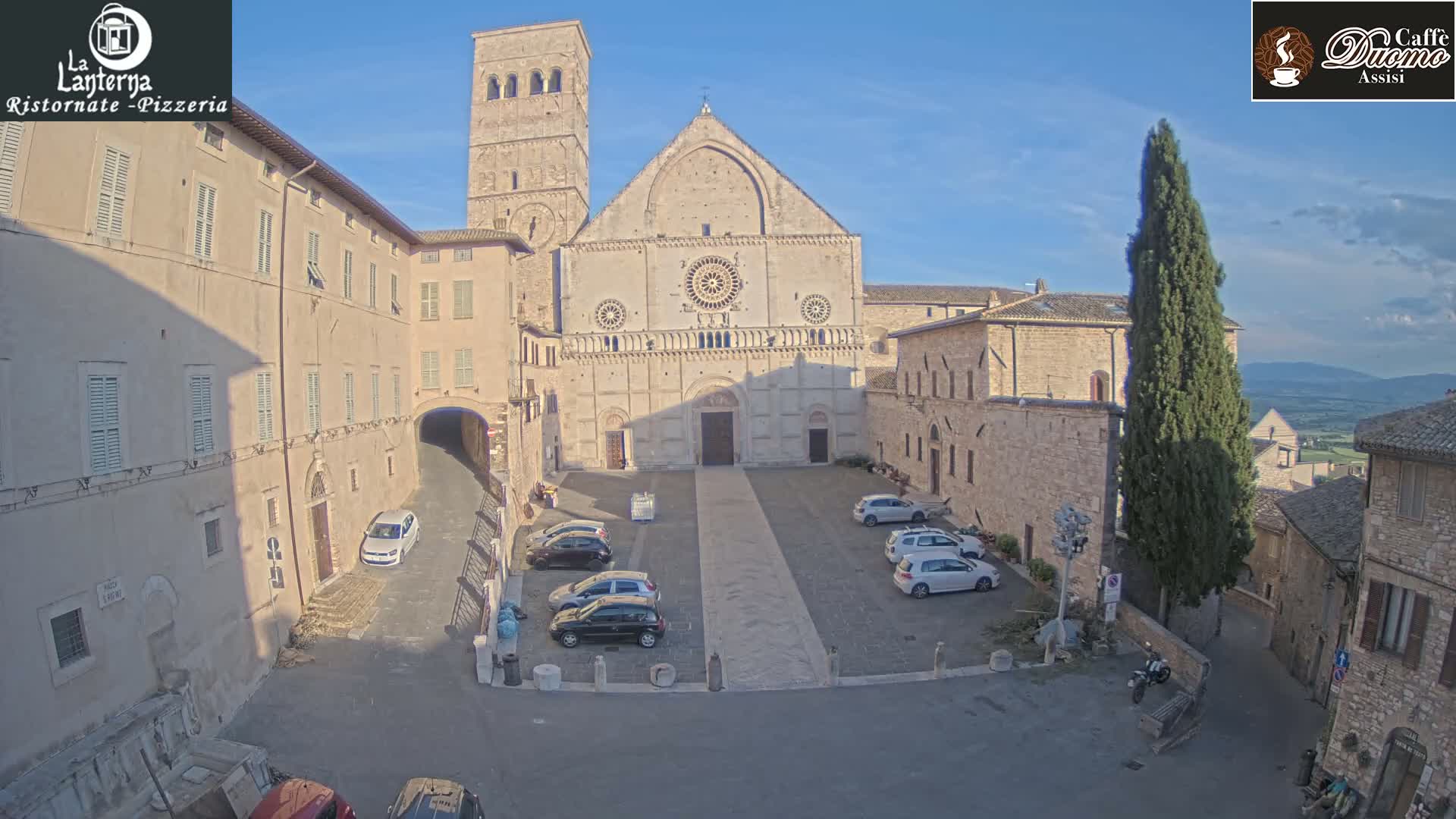 A large stone cathedral sits in a town square under a clear blue sky, with several cars parked nearby and buildings surrounding the square.