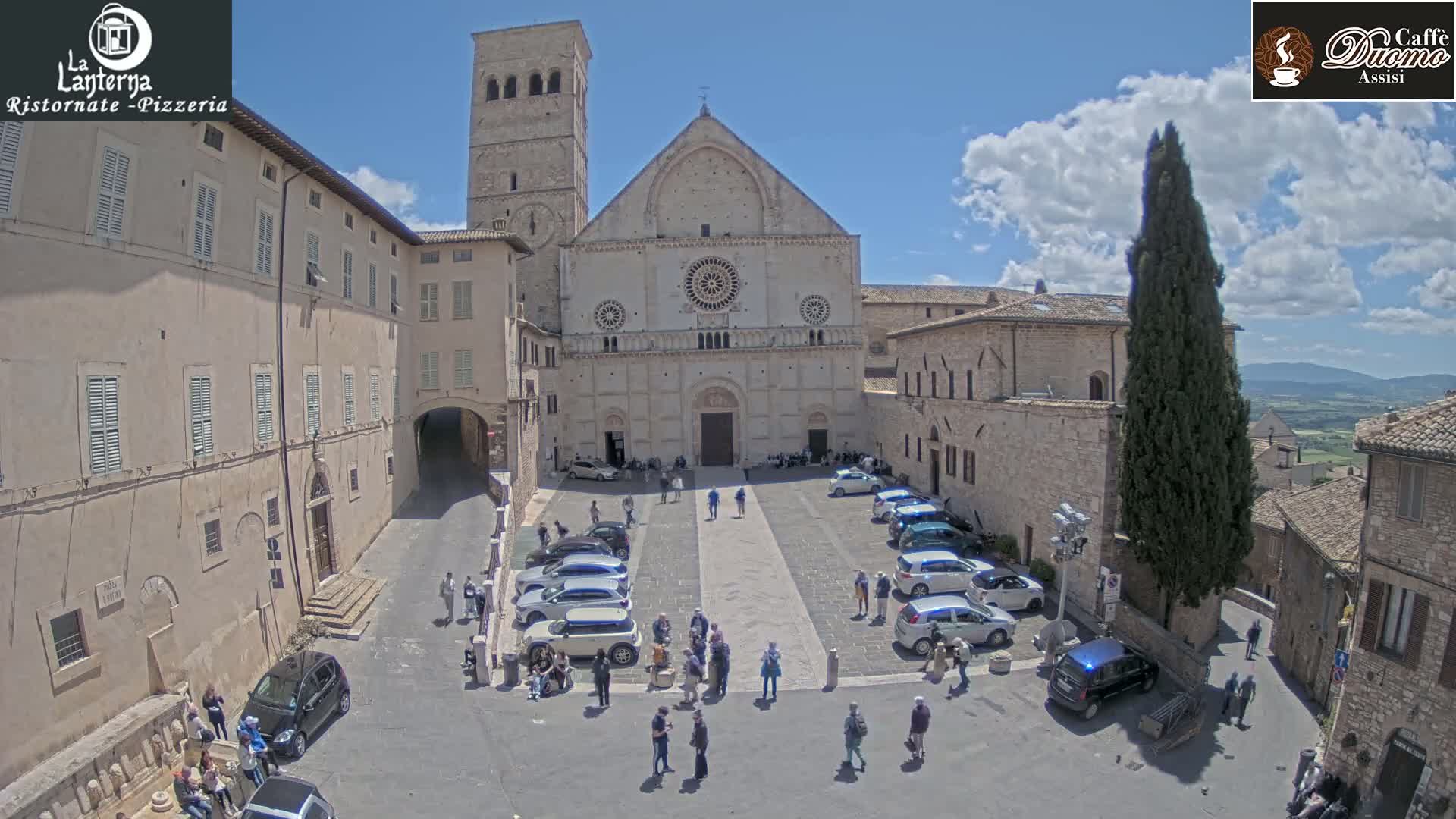 A sunny day reveals a town square with a large stone cathedral at its center, surrounded by parked cars and pedestrians, and adjacent buildings.