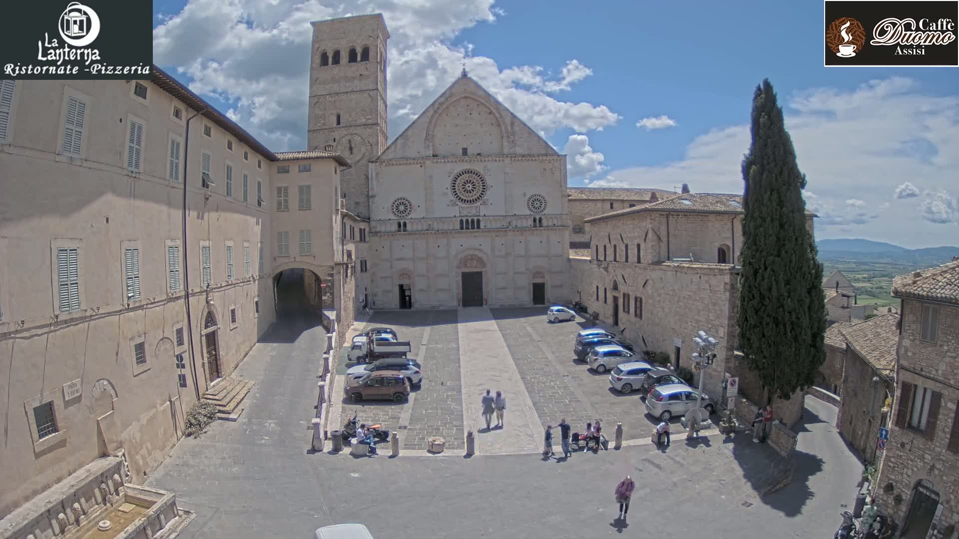 A sunny, partly cloudy day reveals a large stone plaza with parked cars, pedestrians, and a grand cathedral at its center, flanked by older stone buildings.